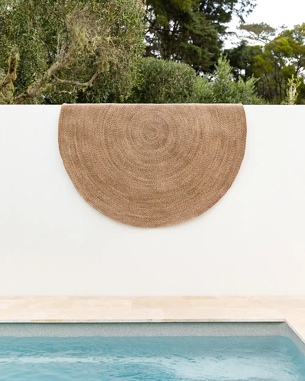 A round, brown rug by Baya NZ hangs over a wall by a swimming pool.