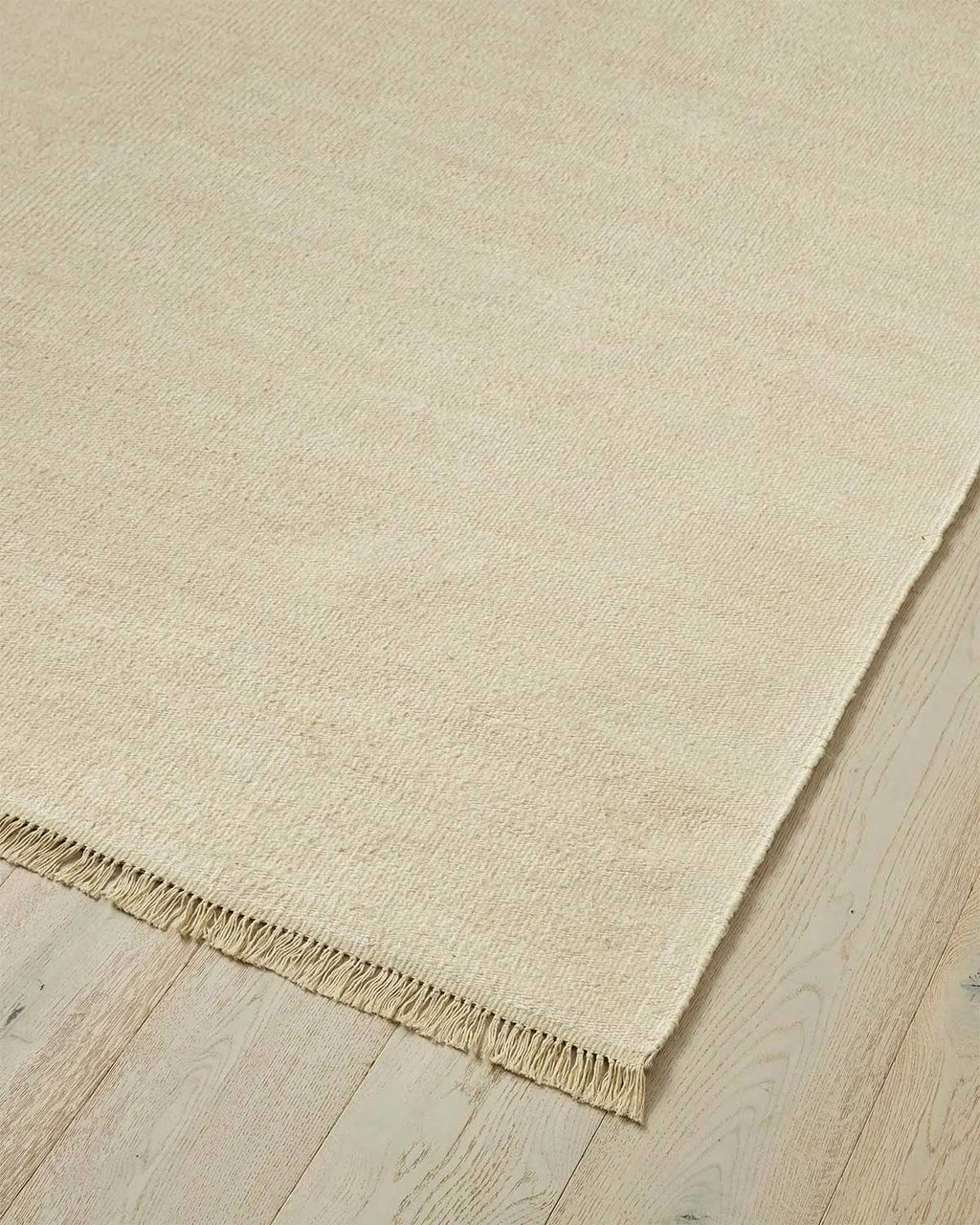 A corner of a 100% linen fibre floor rug in a soft warm cream tone with a matching fringed edge.