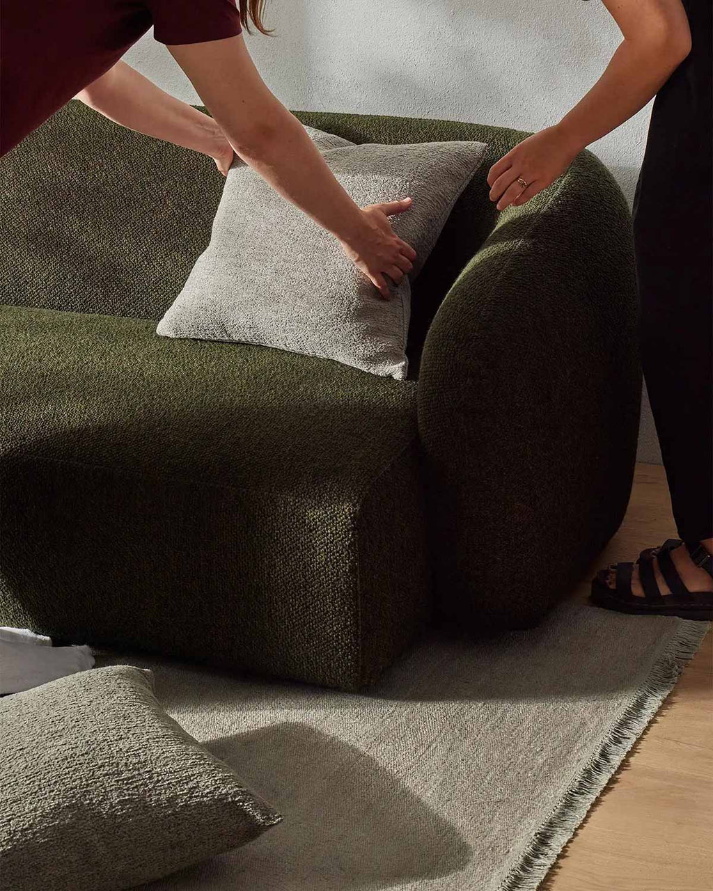 Two people placing a cushion on a modern style green couch, in a room with a cool beige toned floor rug in linen fibres.