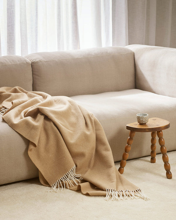 A gorgeous sand toned neutral throw blanket in 100% merino wool draped over a cream couch.
