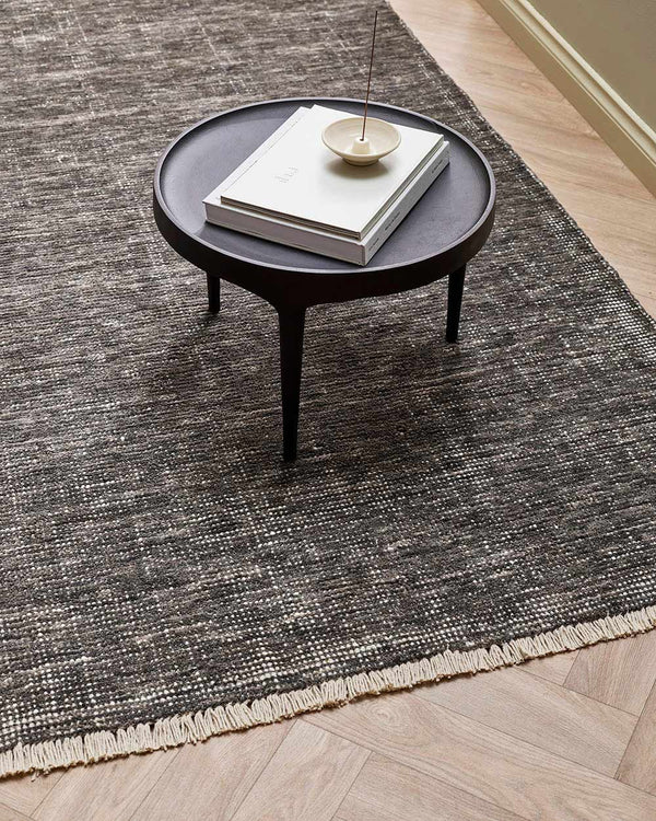 An edgy, slightly distressed 100% wool floor rug in a charcoal peat tone, with white detailing including a subtle fringed edge.