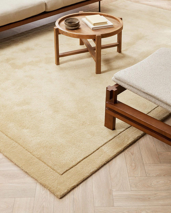 A floor rug with modern furniture in warm soft beige tones with cut pile frame detailing along the edges, in 100% NZ wool.