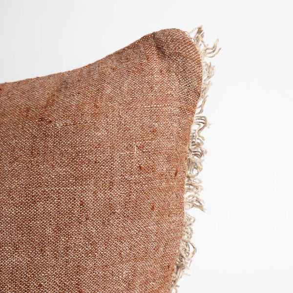 The corner of a warm, tobacco-toned cushion with cream fringing along the edges.