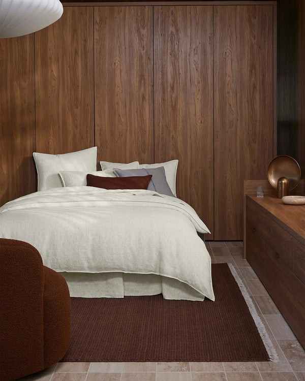 Modern bedroom with wooden wardrobe and bed with white bedding, and a warm clay brown floor rug with white edges.