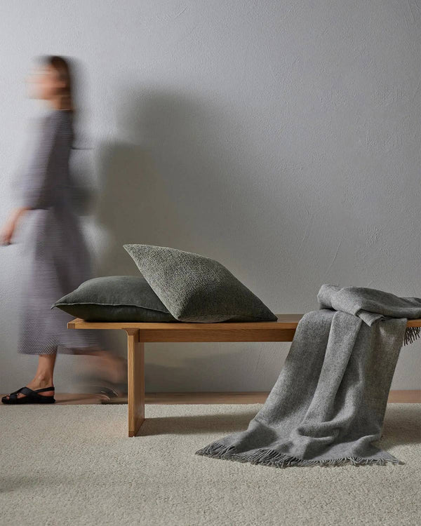 A woman walking through a room with a bench containing grey cushions and a slate-toned throw blanket, which contrasts nicely with the off-white loop pile floor rug.
