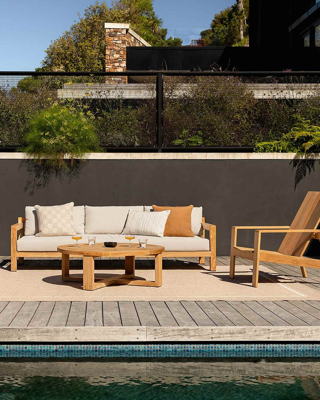 A couch and deckchair by a swimming pool on a sunny day, with a woven outdoor rug by Baya NZ