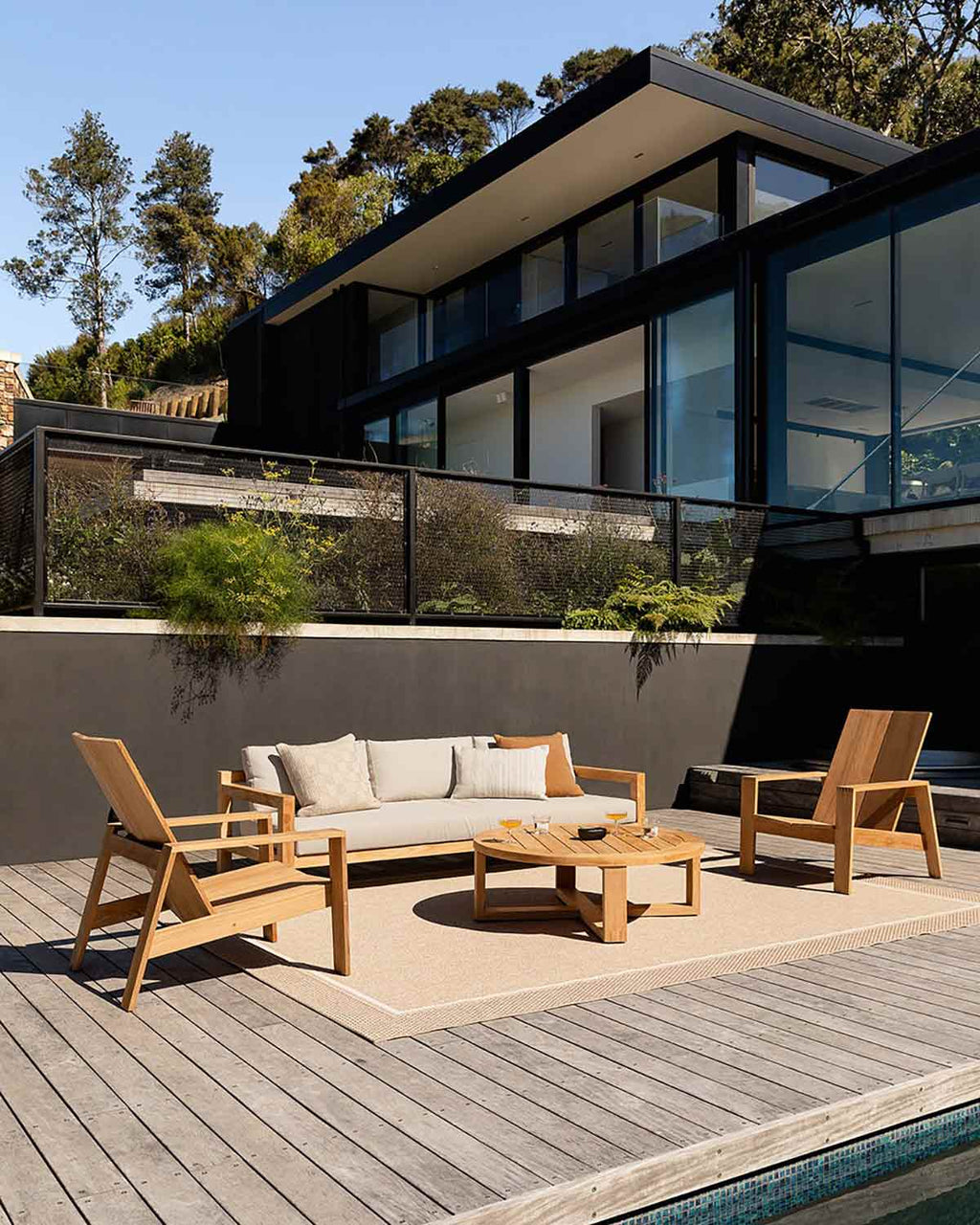 A couch and deckchair by a swimming pool on a sunny day, underneath a modern architectural house with a woven outdoor rug by Baya NZ