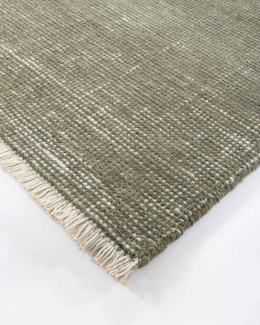 A corner image of the Corbett floor rug in Lichen by Baya, with light distressing in sage green and a short cream fringe.