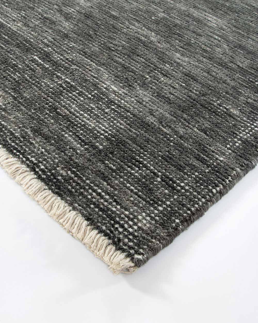 A corner image of the Corbett floor rug, in shade Peat, a soft charcoal with contemporary distressing with a short cream fringe. By Baya