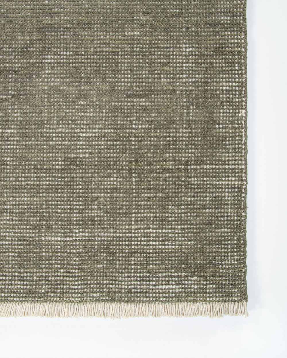 A corner image from above of the Corbett floor rug in the shade Lichen, a lightly distressed contemporary sage green, with cream detailing and a short creamy fringe. By Baya