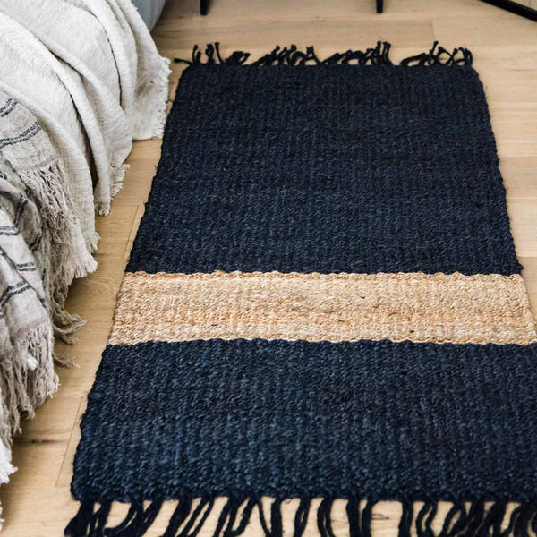 A floor mat next to a bed, in graphite/black jute fibres with a bold natural stripe and fringed edges.