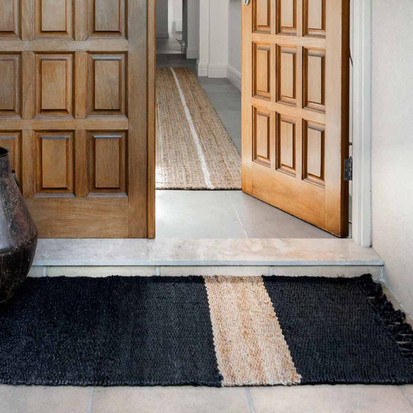 A doorway with a large black jute entrance mat with a natural brown stripe and fringed edges.