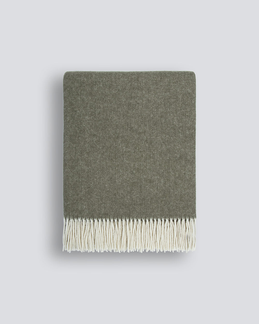 A neatly folded muted olive-green 100% merino wool throw blanket with a soft texture and a delicate cream fringe along the edge, by Baya