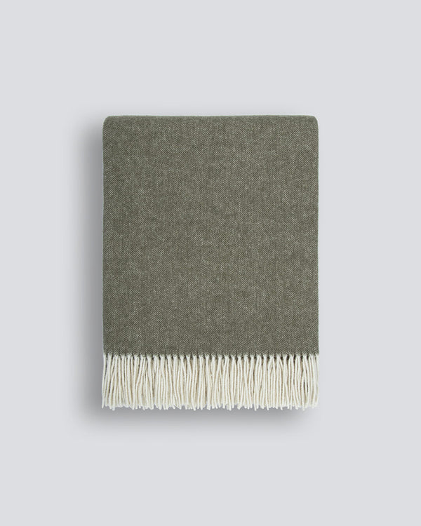 A neatly folded muted olive-green 100% merino wool throw blanket with a soft texture and a delicate cream fringe along the edge, by Baya