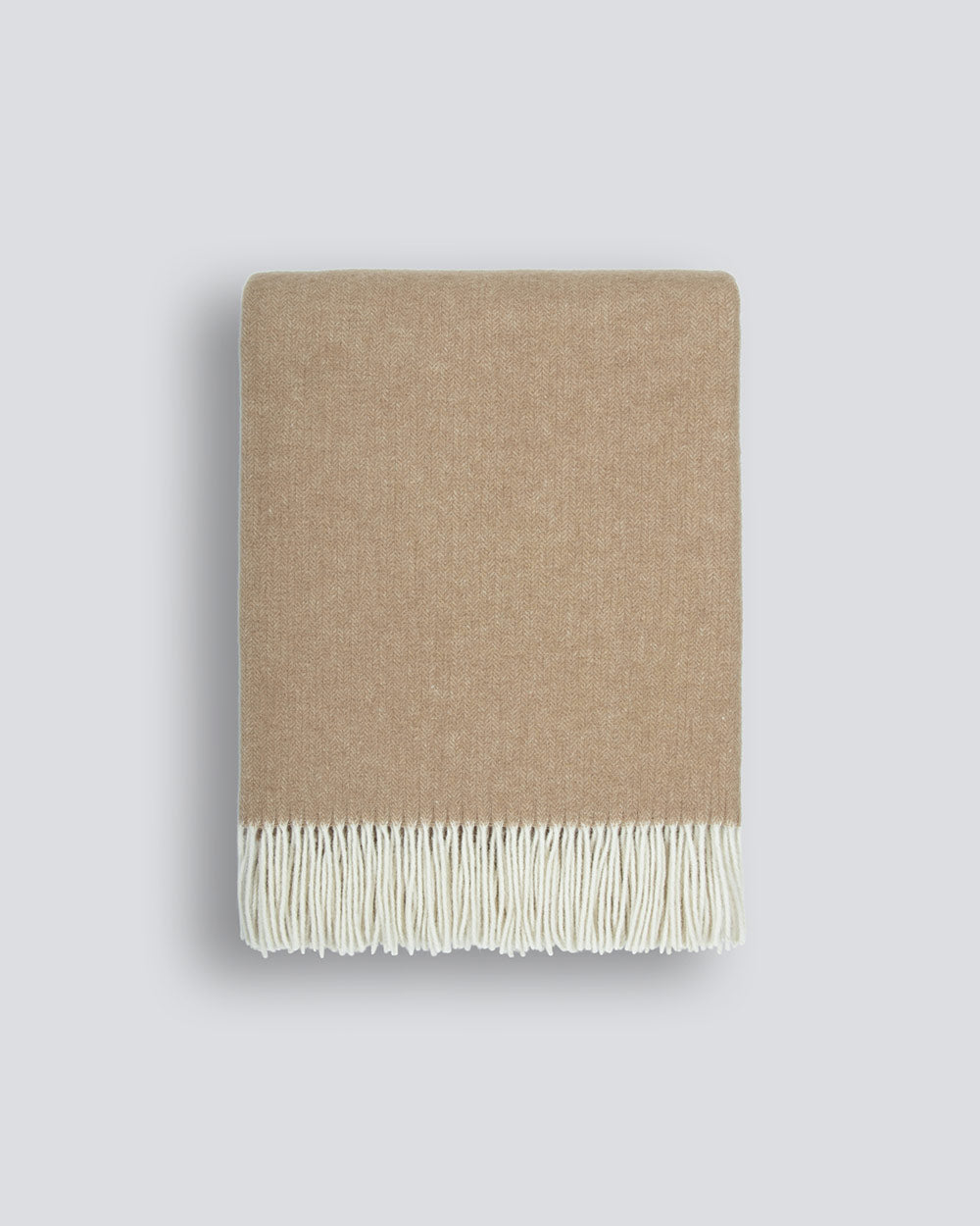 A neatly folded 100% merino wool throw blanket in warm sand tones with a soft texture and a delicate cream fringe along the edge, by Baya