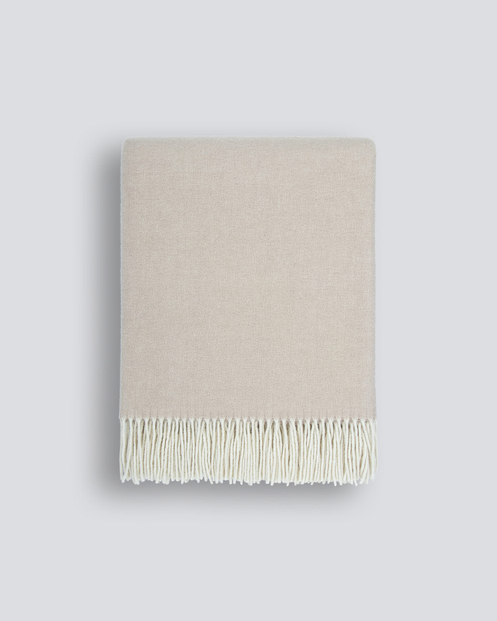 A neatly folded beige 100% merino wool throw blanket with a soft texture and a delicate cream fringe along the edge, by Baya
