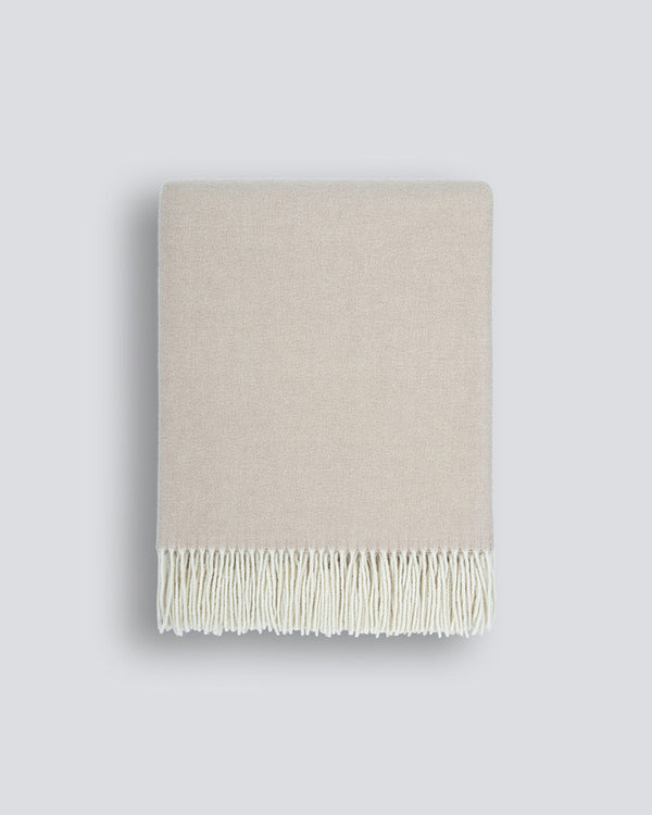 A neatly folded beige 100% merino wool throw blanket with a soft texture and a delicate cream fringe along the edge, by Baya