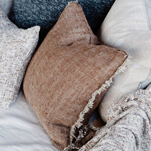 A neutral warm brown cushion with cream fringe detailing across two sides, sitting on a couch.