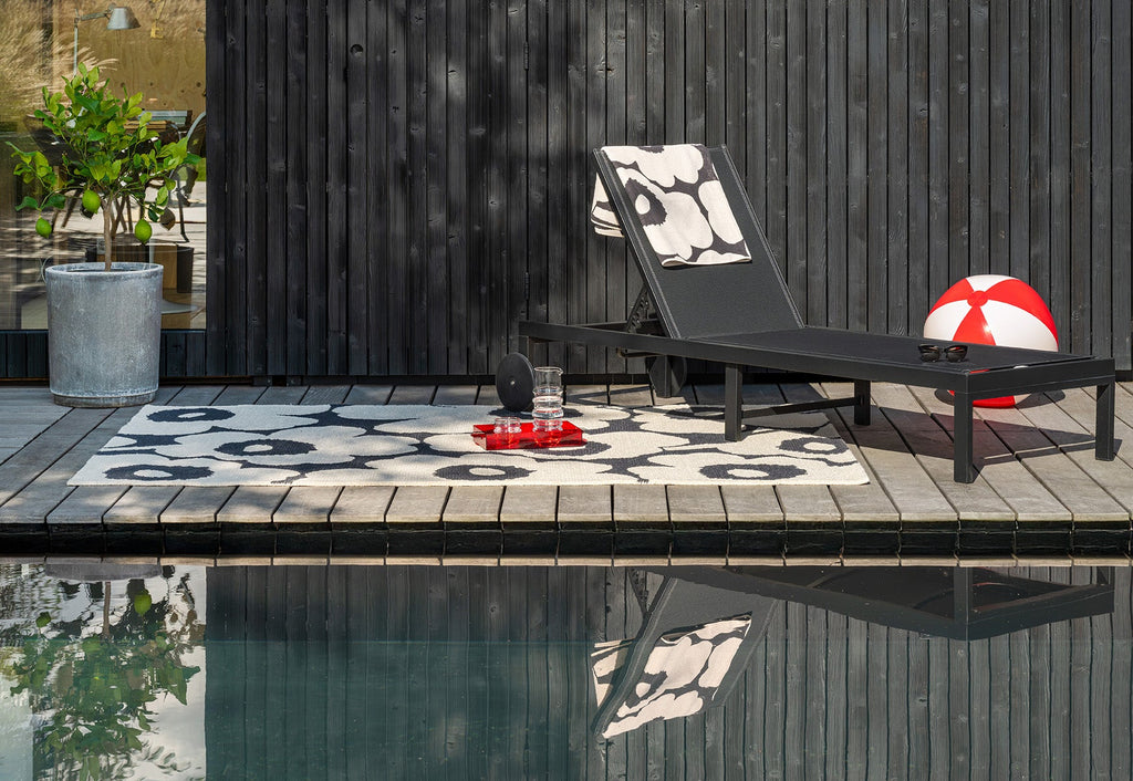 Black lounge chair with a Marimekko Unikko outdoor rug in beige nad black on a wooden deck by a pool.