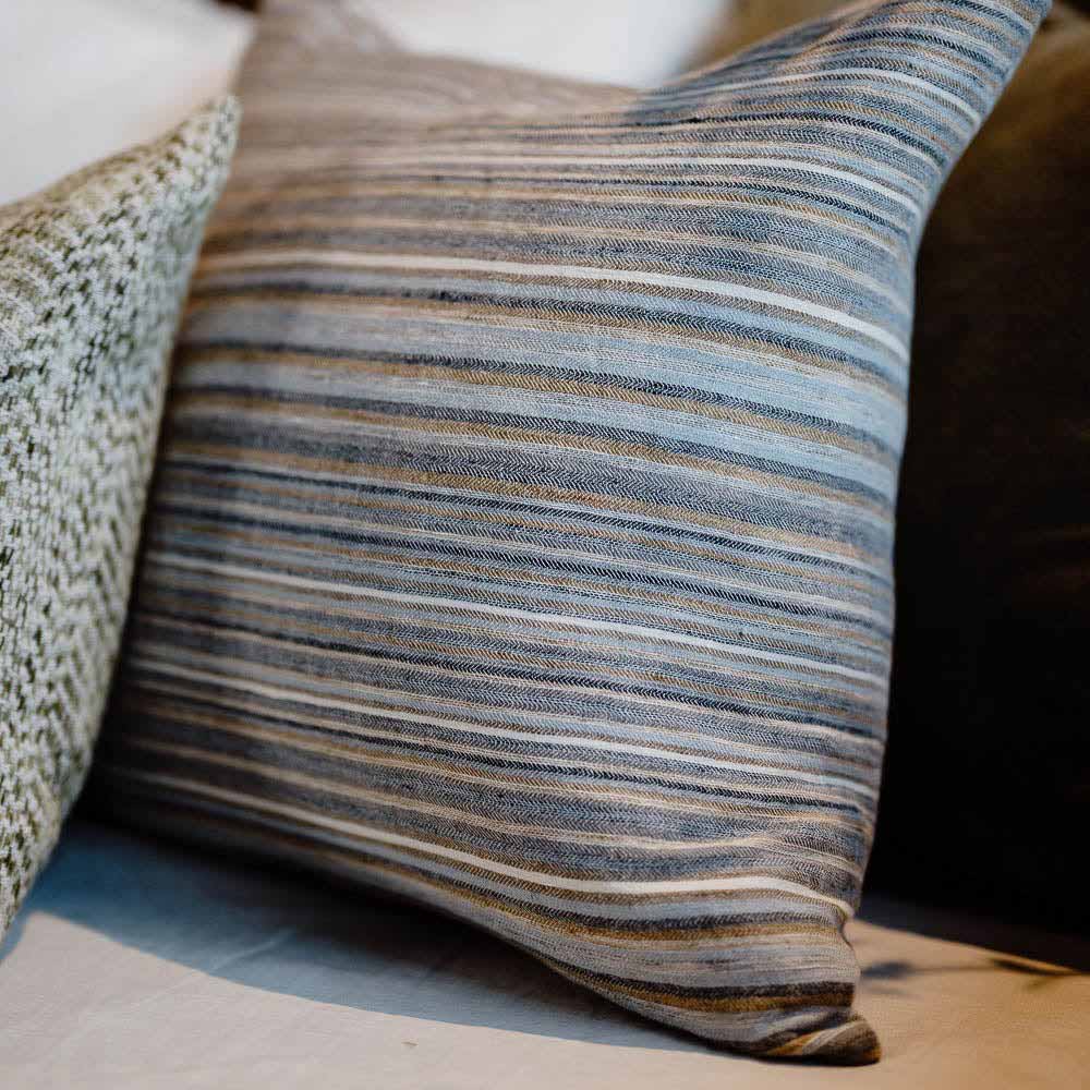 A close up view of the striped cushion in Coast by Eadie. The stripes are irregularly sized, in different tones of muted grey, blue, slate, ochre, and sienna. 