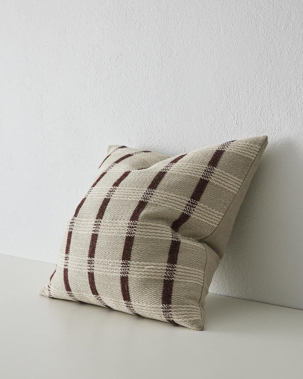 The Weave Home 'Marmaris' woven cushion featuring a dark brown stripe set against a contrasting white and neutrals, seen at three quarter perspective.