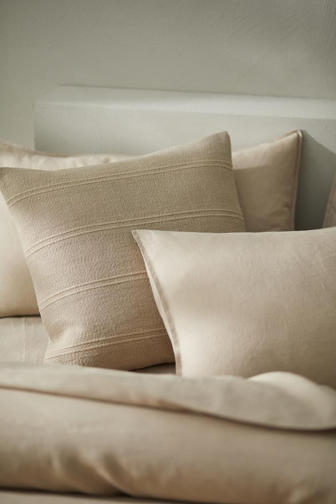 The Weave Home 'Palma' cushion in colour 'Shell', a warm beige neutral, seen on a sophisticated bed setting