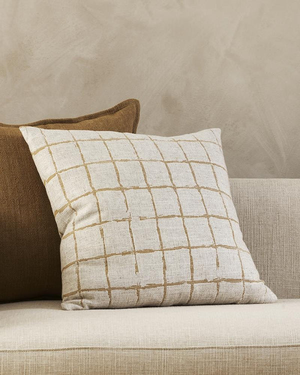 A natural linen-blend cushion with an ochre grid-look design in a hand-painted style, styled with a plain brown cushion.