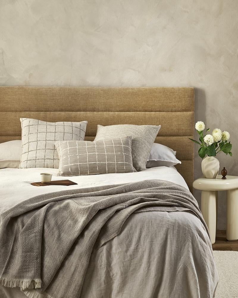 A taupe coloured lumbar cushion featuring a grid design in white in a hand-painted style, the Johnson Taupe is seen here on a bed in a stylish bedroom with other cushions.