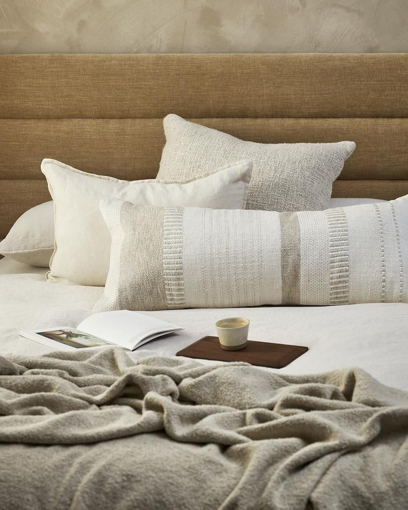 An extra-long lumbar cushion in white and beige - the Bettie by Baya -seen on a bed with cream and white cushions.