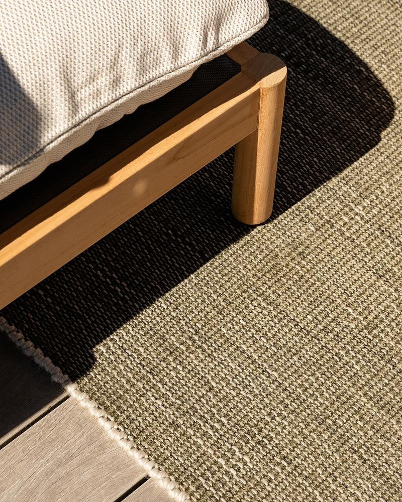 The olive green handwoven Lonsdale outdoor rug by Baya, seen inthe sun under outdoor furniture