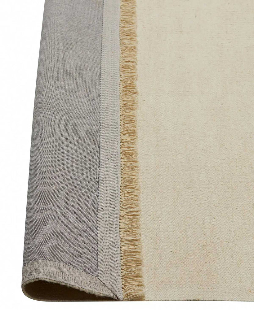 A corner of a linen floor rug in a soft cream tone with the edge flipped up to show the durable cotton backing and the underside of the fringed edge. 