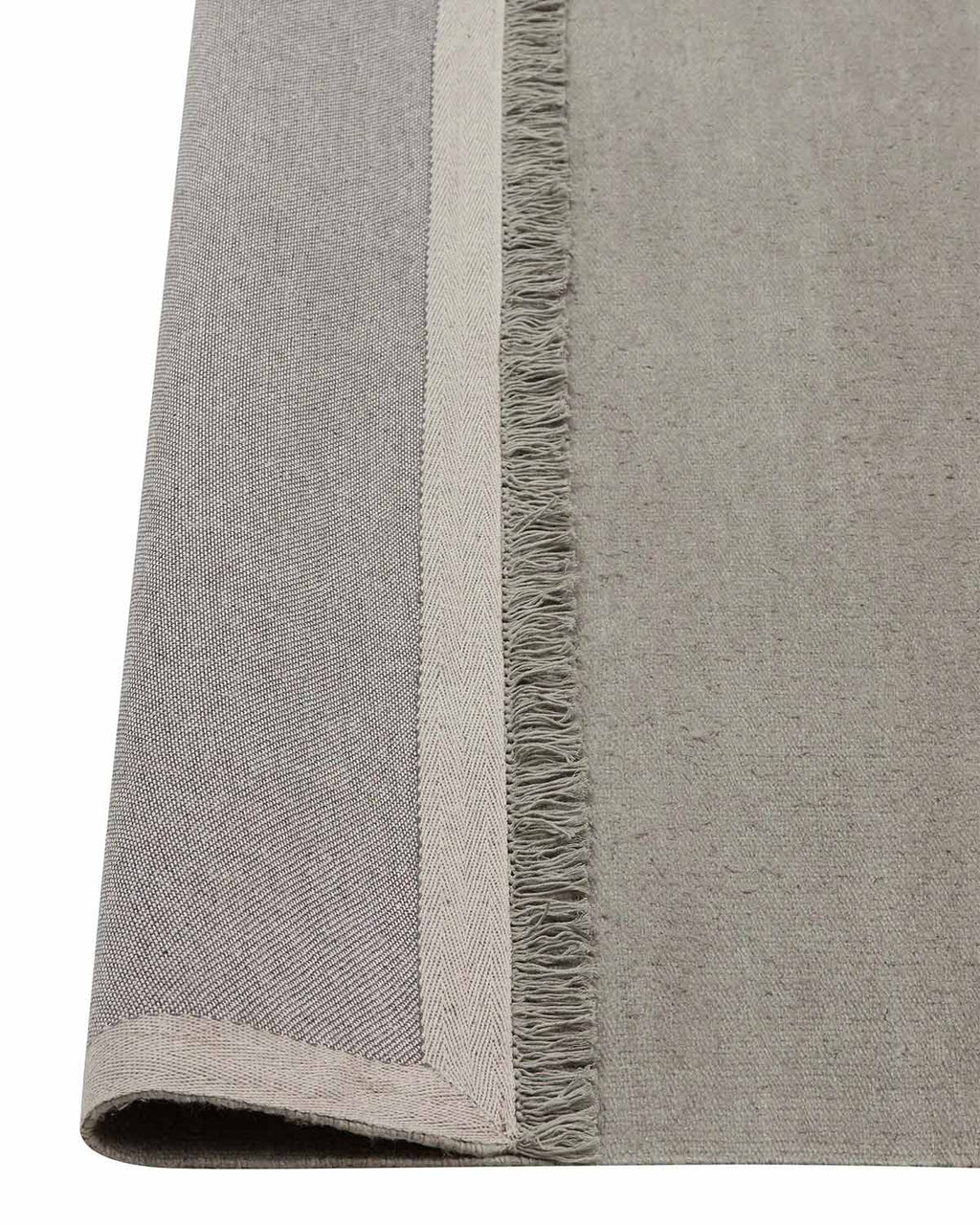 A corner image of a lichen toned grey green floor rug in linen with the edge flipped up to show the underside of the fringed edging and the durable cotton backing. 