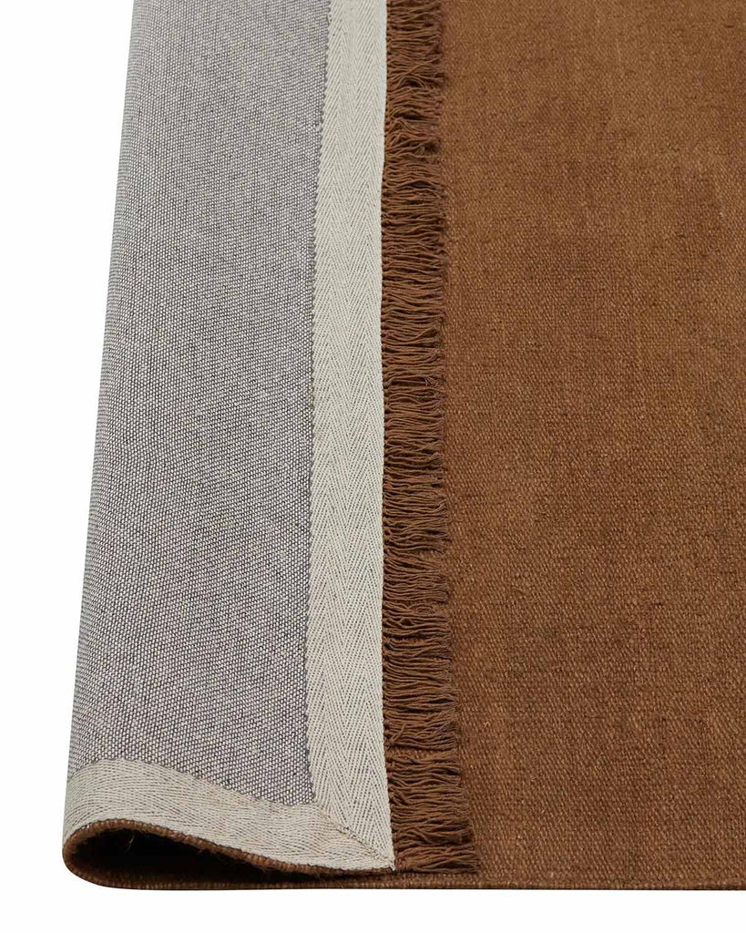 A corner image of a warm brown floor rug made with linen fibres with the edge flipped up to show the underside of the fringe and the durable cotton backing. 