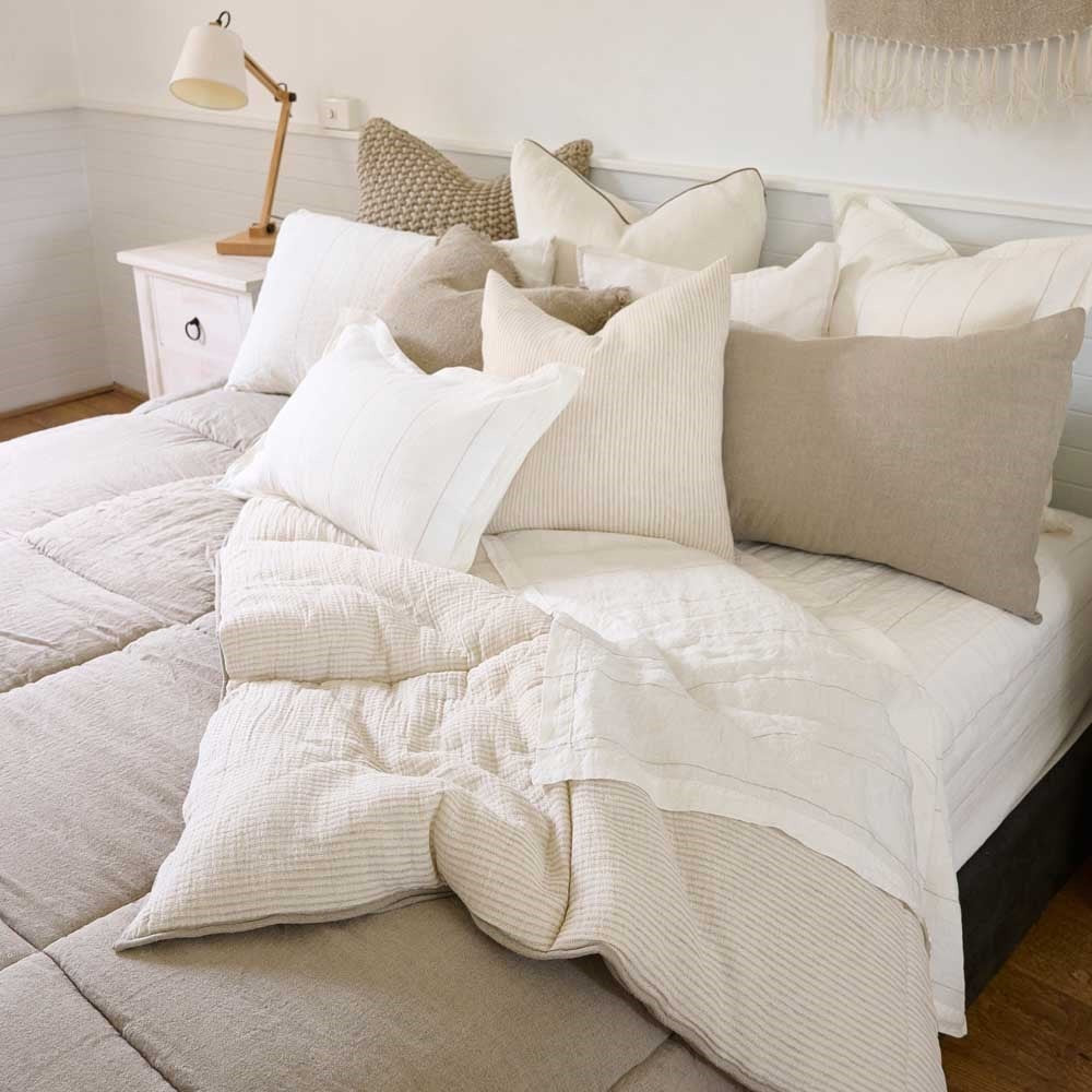 A very cosy looking bed with a number of cushions in neutral creams and taupes, and a quilted bedcover with a reversible beige pinstripe inner. 