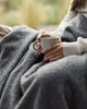 Close up of the Weave Home wool throw blanket in colour 'charcoal' keeping a lady warm while she drinks coffee