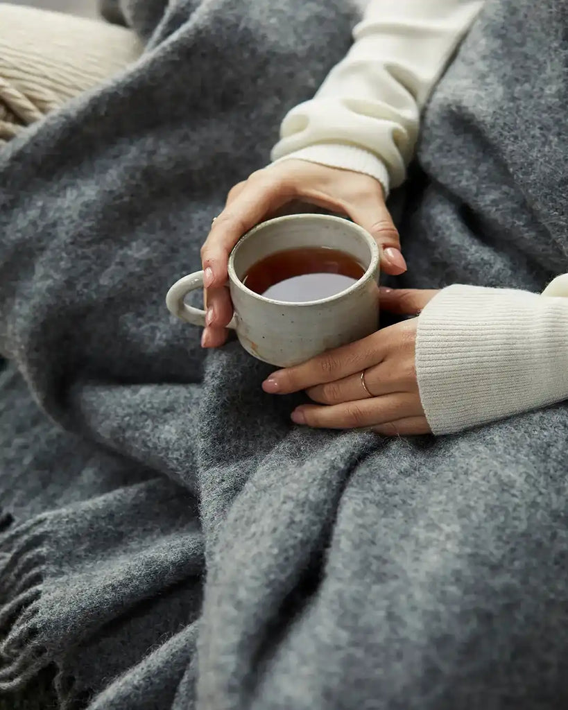 Close up of the Weave Home wool throw blanket in colour 'charcoal' on a lady's lap while she drinks coffee