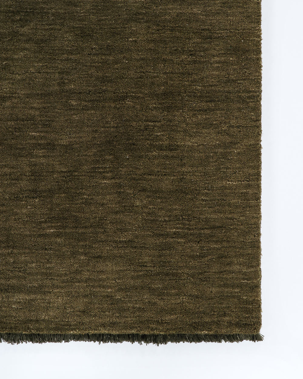 A corner image seen from above of a warm rich green floor rug in 100% wool with fringed edges. 