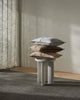 Stack of pillows on a small round table against a neutral wall with curtains.