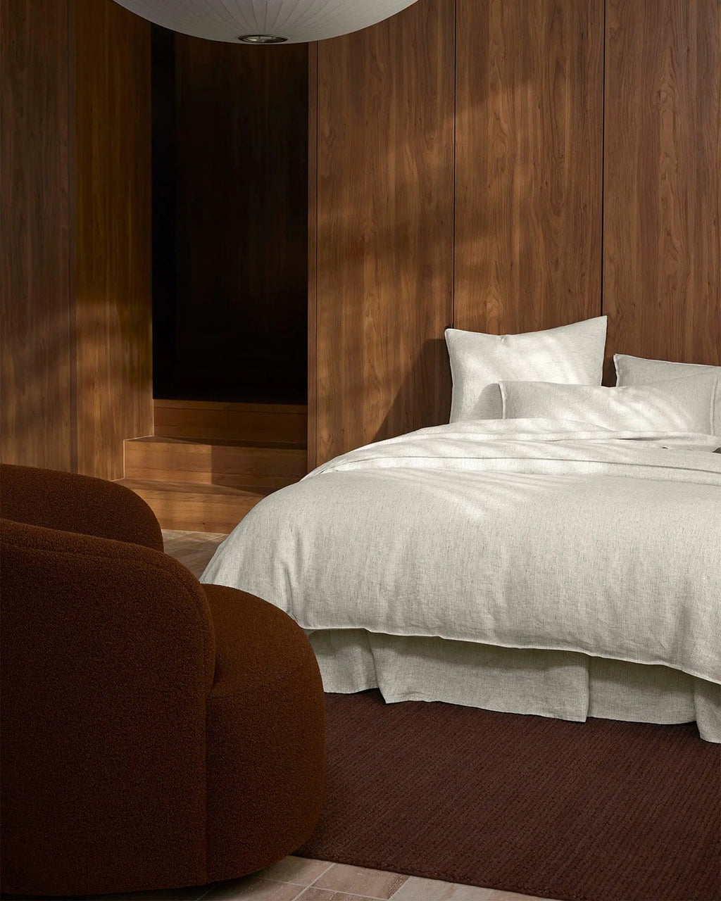 Modern bedroom with wooden wall, bed, and brown armchair, as well as a deep warm toned clay-brown rug. 