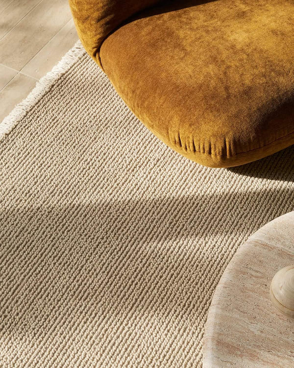 Close-up of a textured beige rug with a brown ottoman partially visible.