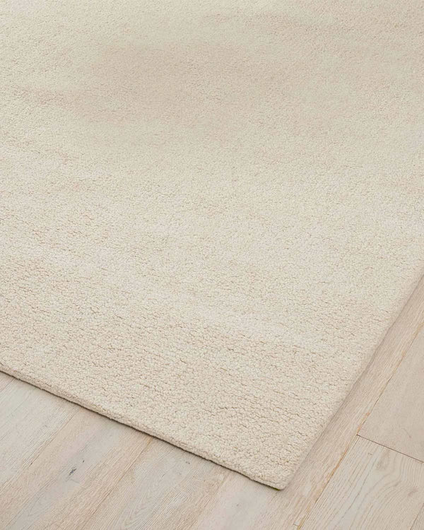 A corner image of a wool blend rug in a soft cream tone with a textured pile. 