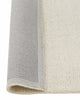 A corner image of a loop pile floor rug in light soft cream tones with the edge flipped to show the durable cotton backing. 
