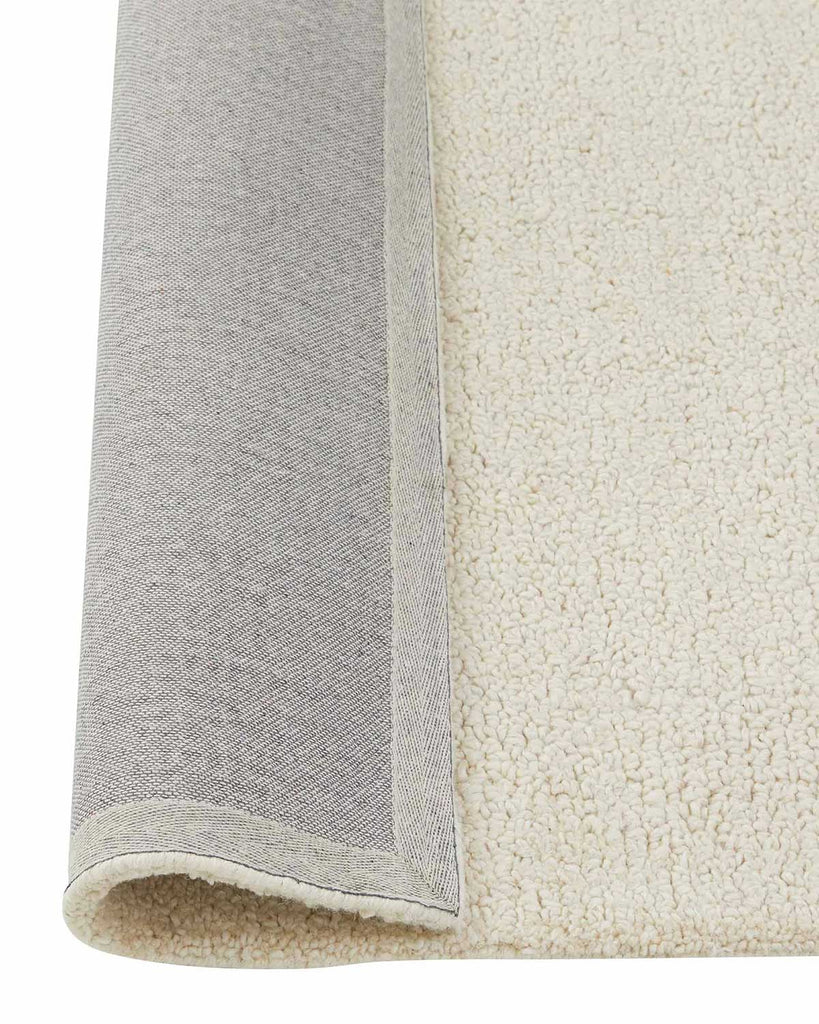 A corner image of a loop pile floor rug in light soft cream tones with the edge flipped to show the durable cotton backing. 