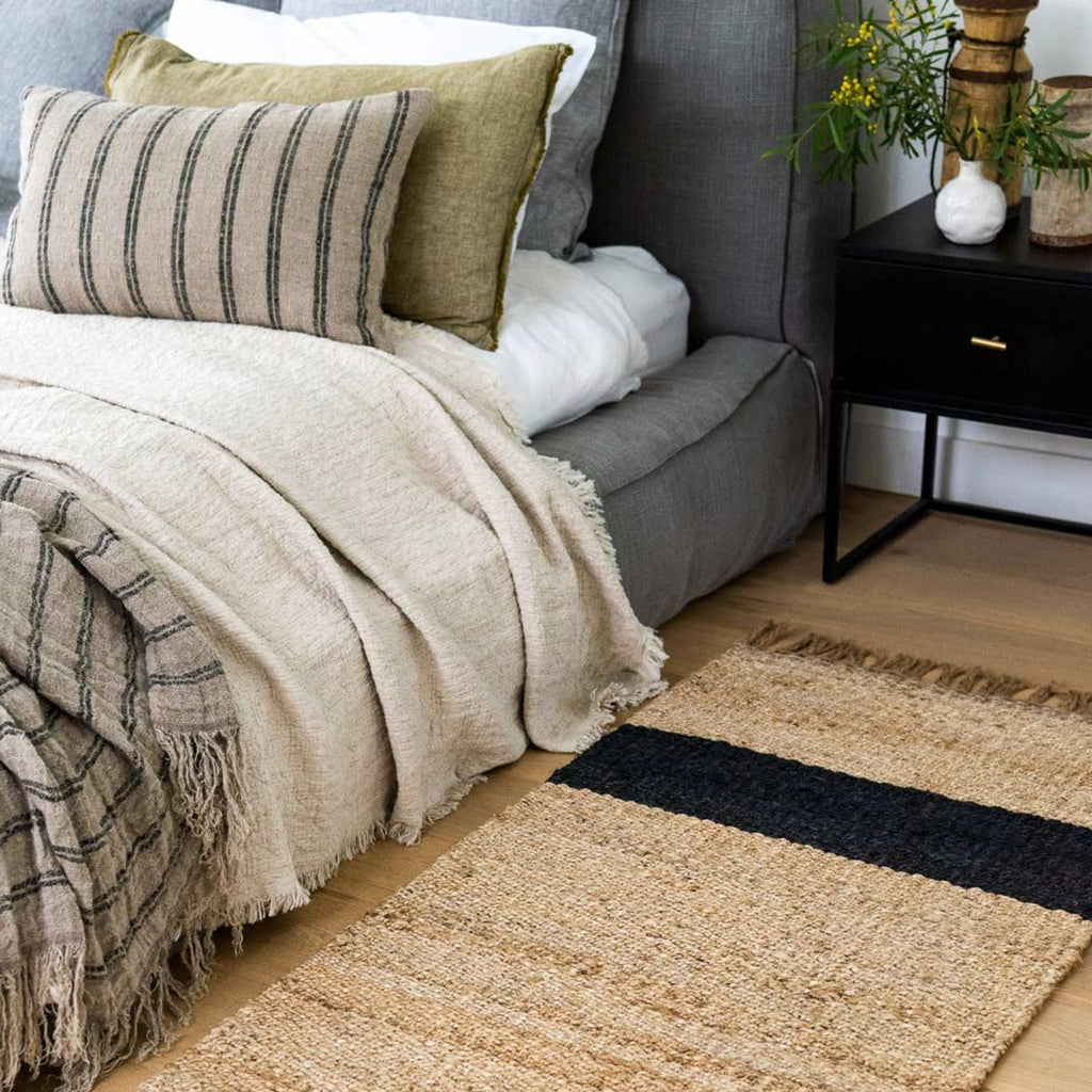 A bedroom with a jute mat by the bed, in natural brown tones and organic fibres, with a bold graphite asymmetrical stripe and fringed edges. 