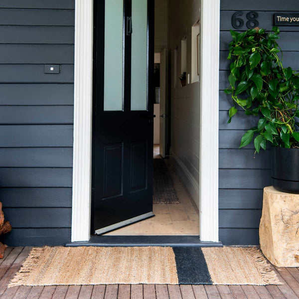 A doorway with an oversized jute entrance mat with fringed edges and an asymmetrical stripe on one side, by Zebra Home. 