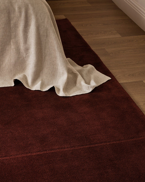Baya Bastion NZ wool floor rug in Claret, close view at the foot of a bed with a linen throw, on a timber floor.