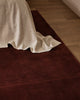 Baya Bastion NZ wool floor rug in Claret, close view at the foot of a bed with a linen throw, on a timber floor.