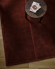 Baya Bastion NZ wool floor rug in Claret, overhead view beneath a small round side table with books, on a timber floor.