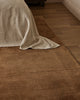Baya Bastion NZ wool floor rug in Hazelnut, close view at the foot of a bed with a linen throw, on a timber floor.