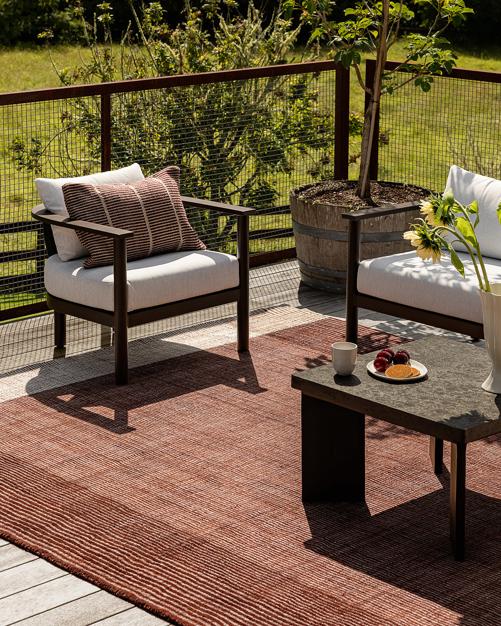 Baya Bedrock Indoor and Outdoor Floor Rug in Claret, styled on an outdoor deck under a timber armchair with a striped cushion, beside a potted plant and a low stone-topped coffee table set with fruit and sunflowers.